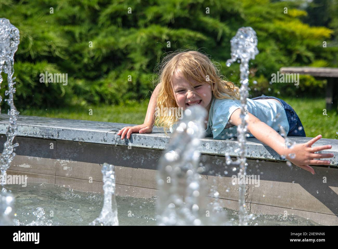 petite fille en jeans et une chemise bleu clair tend la main aux jets d'eau d'une fontaine de ville, ses yeux larges d'émerveillement. Des arbres verts entourent la scène Banque D'Images