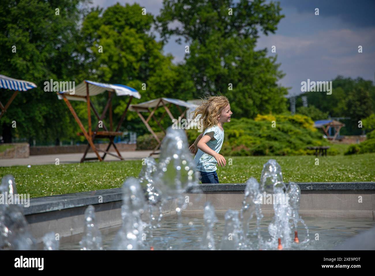 fille en jeans et une chemise bleu clair joue au bord d'une fontaine, l'eau scintillant dans la lumière. Banque D'Images