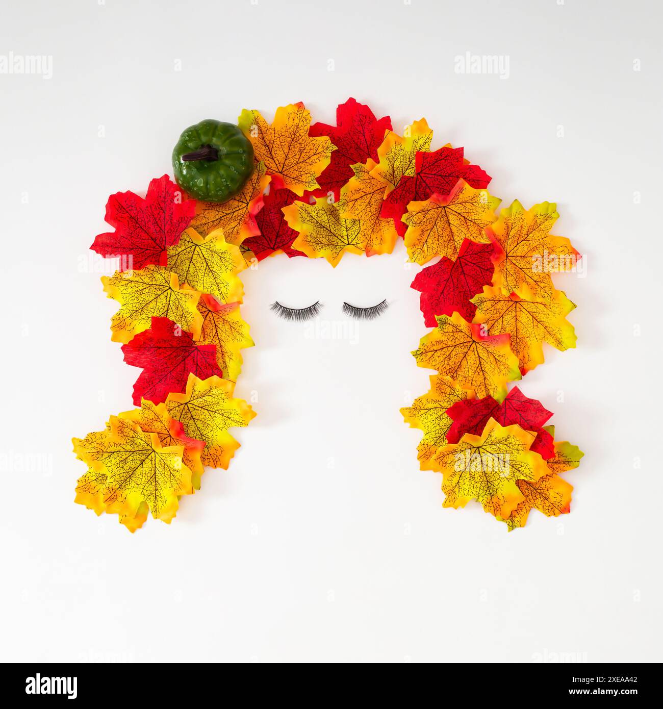 Visage de fille créative fait avec des feuilles d'automne colorées, de la citrouille verte et des faux cils sur fond blanc. Objets d'automne minimaux esthétiques Banque D'Images