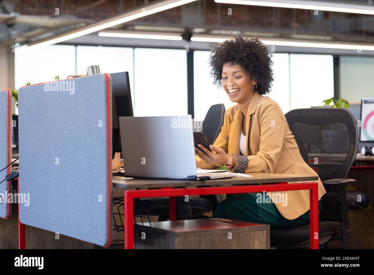 Femme d'affaires afro-américaine utilisant smartphone et ordinateur portable, souriant dans un bureau moderne Banque D'Images