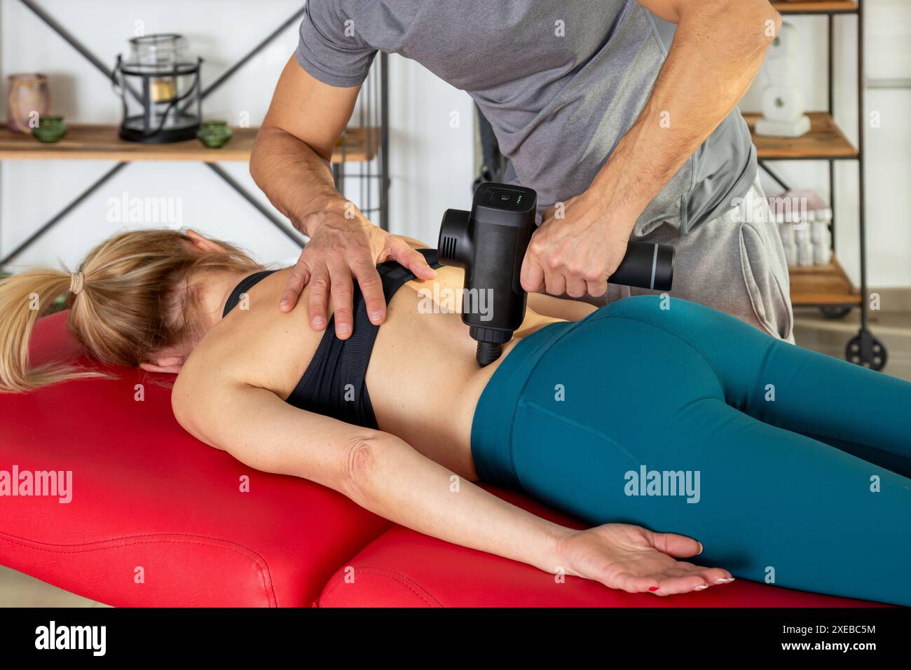 Recadrez l'entraîneur de fitness anonyme en utilisant un pistolet de massage sur le bas du dos de la femme couchée sur le brancard pendant le processus de récupération dans le studio moderne Banque D'Images