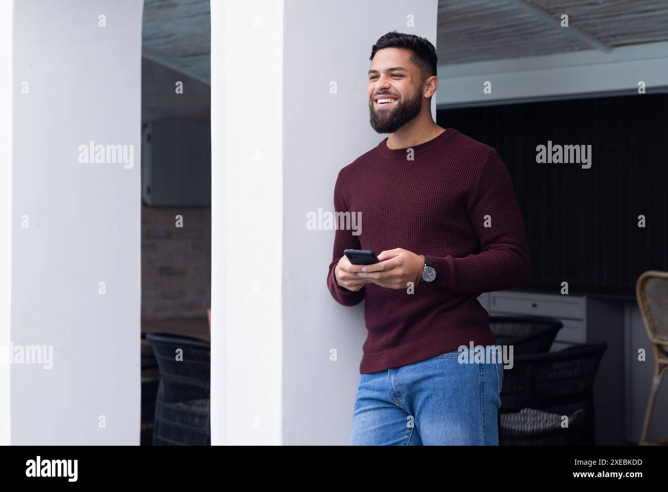 En dehors de sa maison, homme souriant tenant smartphone et appuyé contre pilier à la maison Banque D'Images