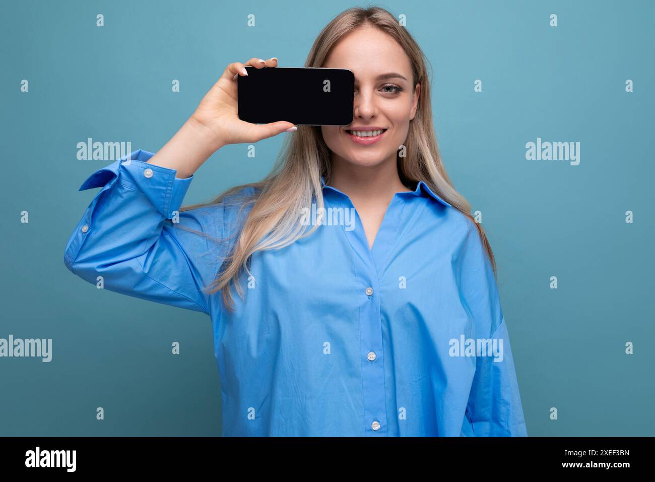 Fille blonde informelle souriante tenant un smartphone avec maquette d'écran vierge pour le site Web sur fond bleu Banque D'Images