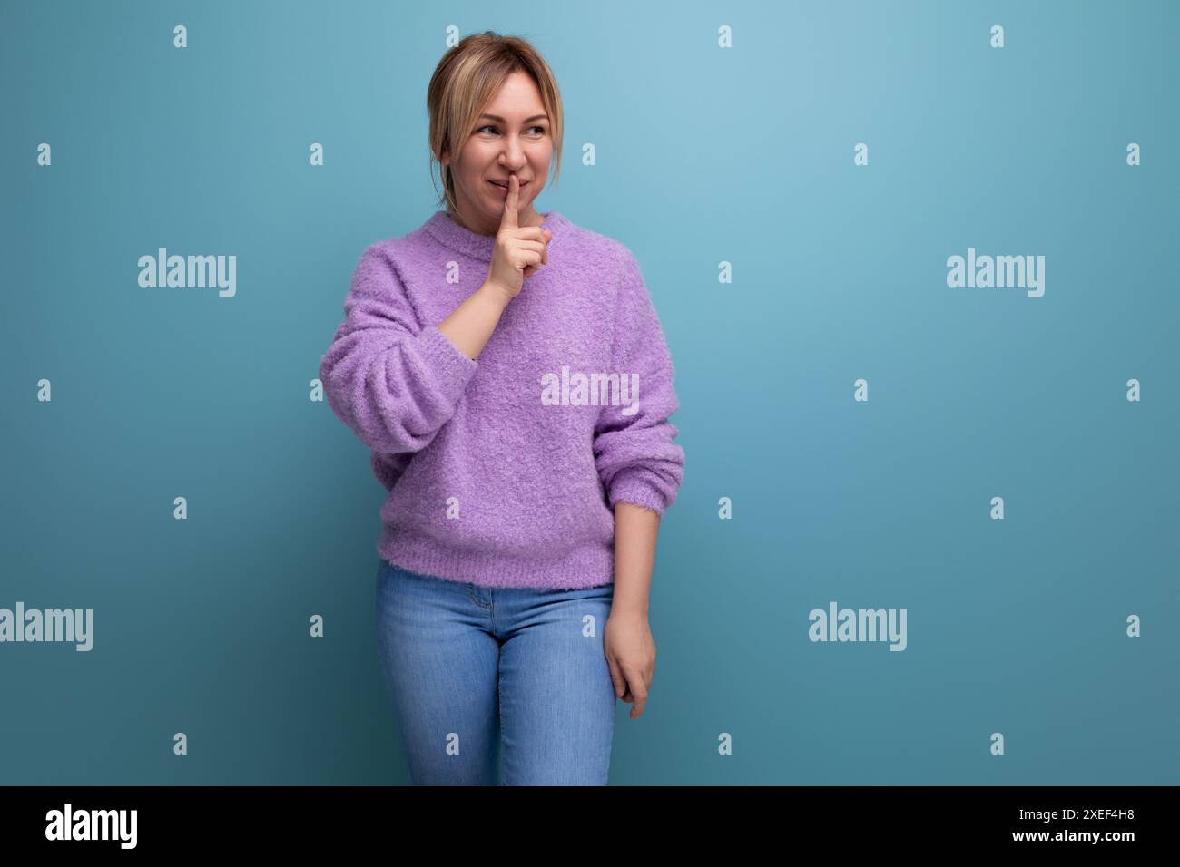 Jeune femme blonde mignonne en sweat à capuche violet montrant un geste de silence sur fond bleu avec espace de copie Banque D'Images