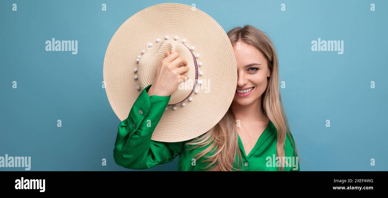 Femme souriante en vacances tenant un chapeau de paille près de son visage sur un fond bleu de studio Banque D'Images