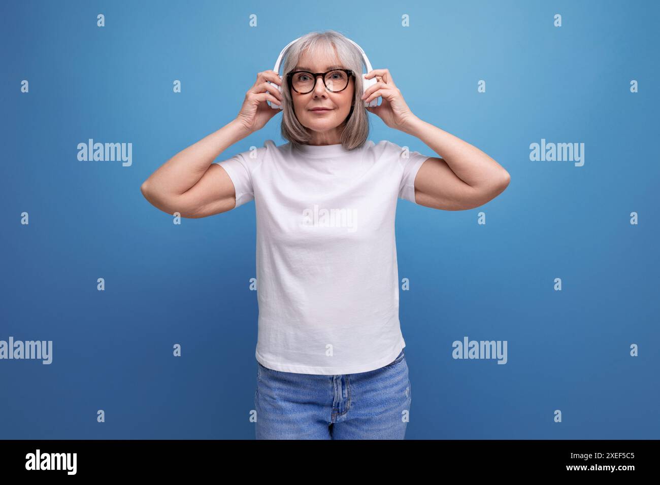Dame mature avec les cheveux gris écoute de la musique dans des écouteurs sans fil sur fond de studio avec espace de copie Banque D'Images