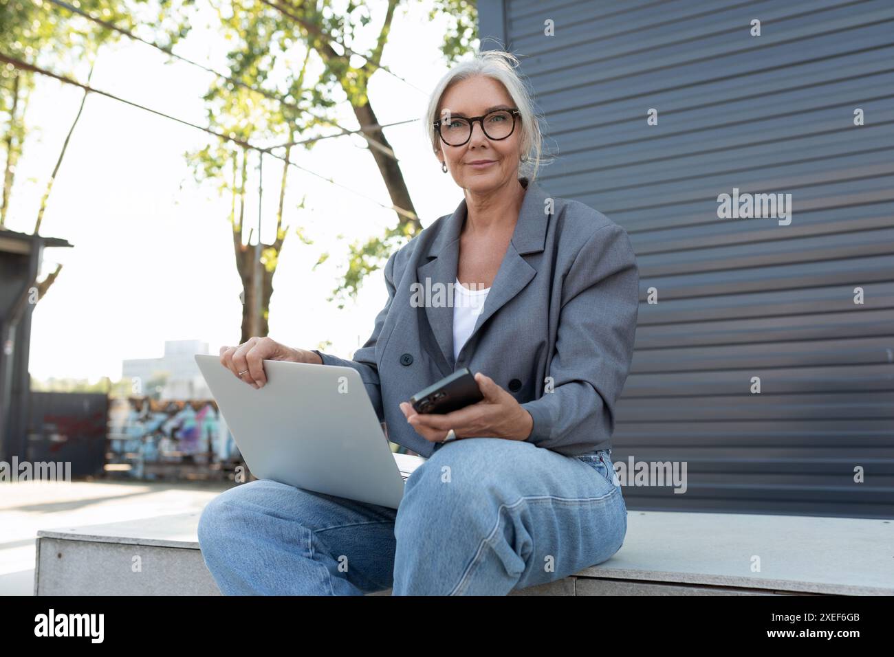 Une jolie femme d'affaires mature aux cheveux gris habillée d'une veste grise élégante et d'un Jean s'est assise sur un banc avec un ordinateur portable Banque D'Images
