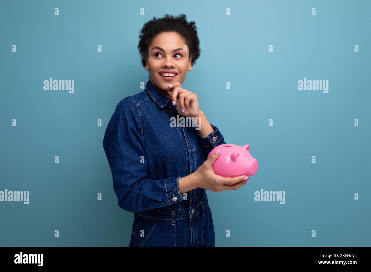 Jeune femme d'affaires hispanique joyeuse avec les cheveux moelleux habillé dans un costume en denim bleu garde des économies dans une tirelire Banque D'Images
