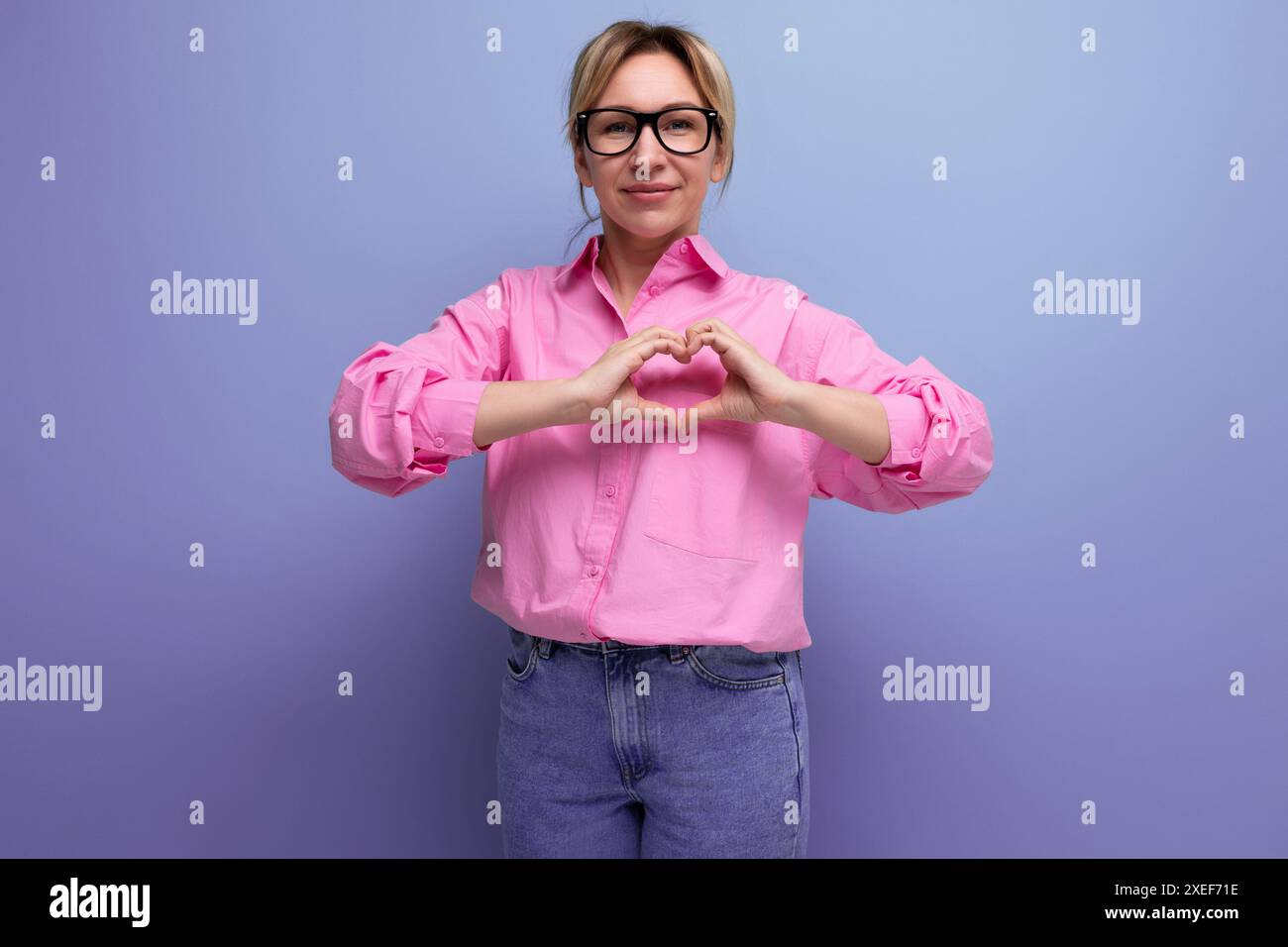 Jeune femme secrétaire blonde caucasienne énergique avec coiffure en queue de cheval, lunettes et dans une chemise rose montre un cœur sur un studio Banque D'Images