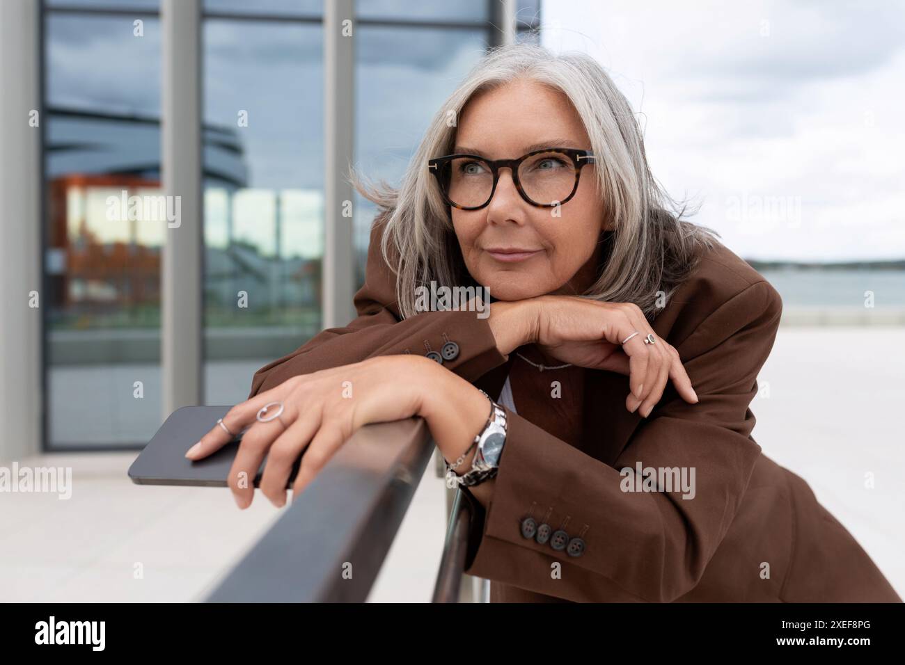 La femme d'affaires mûre aux cheveux gris mince réussie est habillée dans une veste brune élégante sur le fond du bâtiment Banque D'Images