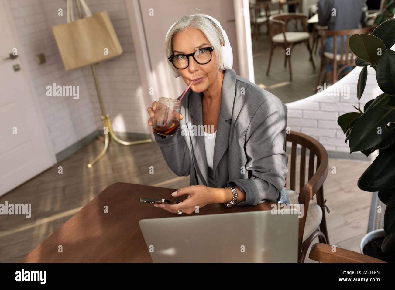 Une jolie femme d'affaires mature aux cheveux gris habillée d'une veste grise élégante est assise dans un café et regarde une vidéo sur un ordinateur portable Banque D'Images