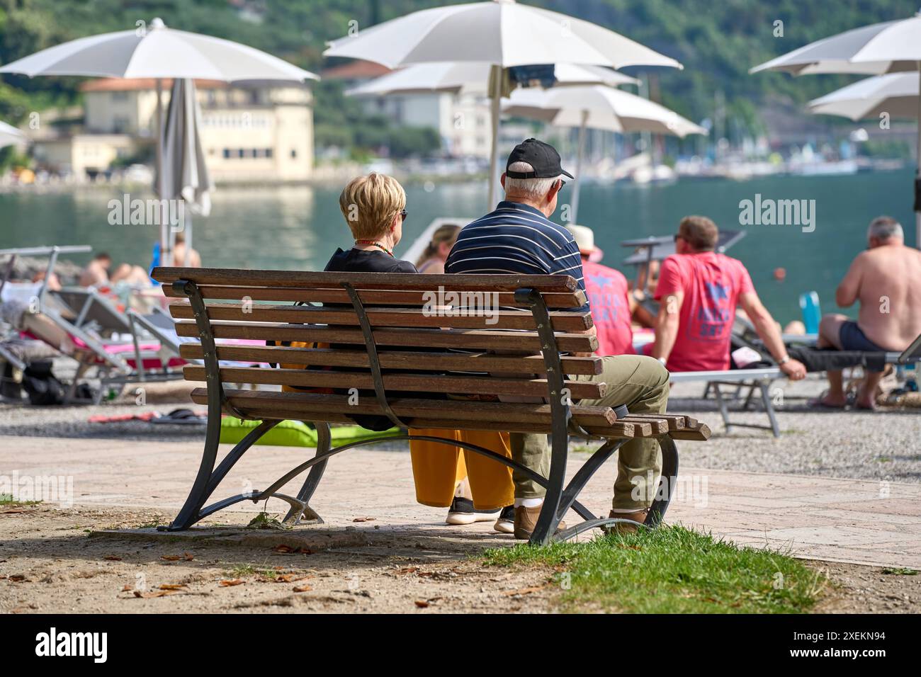 Nago Torbole, Lac de Garde, Italie - 27 juin 2024: personnes âgées profitant d'un moment de détente sur un banc de parc sur la rive du lac de Garde, entourées d'autres touristes et amateurs de plage sous des parapluies *** Senioren genießen eine entspannte Zeit auf einer Parkbank am Seeufer am Gardasee, umgeben von anderen Touristen und Strandbesuchern unter Sonnenschirmen Banque D'Images