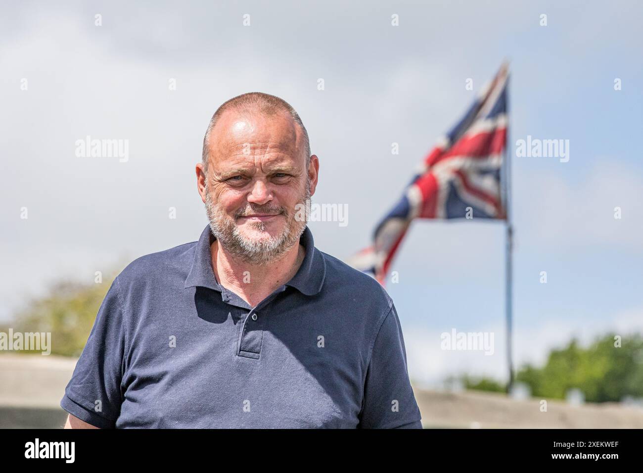 Le comédien Al Murray dehors au soleil vêtu d'un polo bleu avec l'Union Jack volant sur un mât de drapeau en arrière-plan Banque D'Images