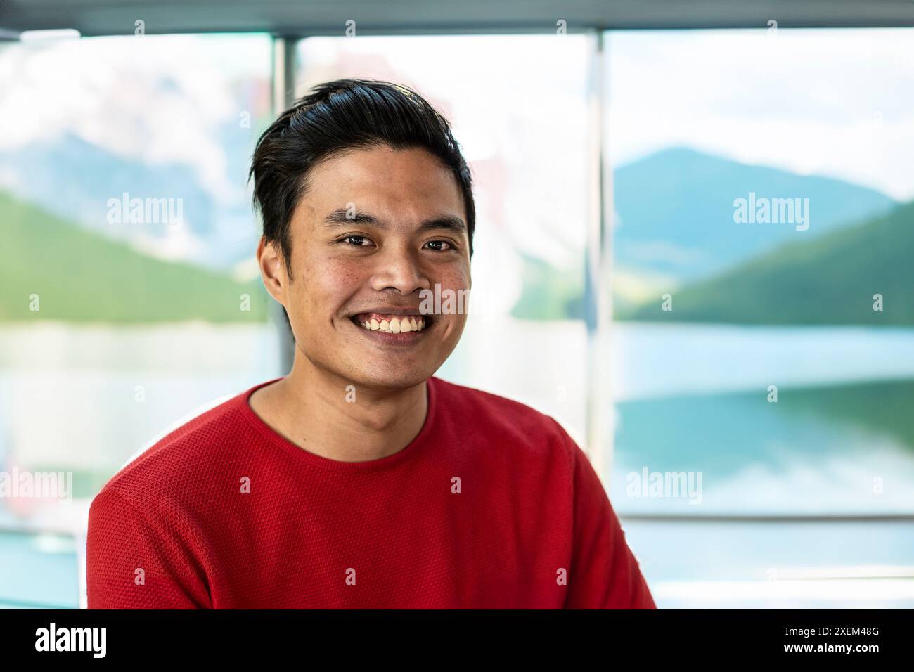 Jeune homme asiatique souriant à l'intérieur avec une vue panoramique sur la montagne et le lac en arrière-plan. Banque D'Images