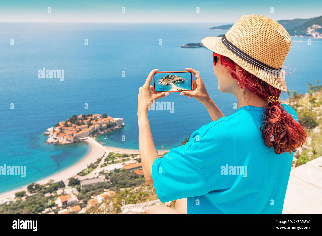 Une femme voyageuse se tient au point de vue de l'île de Sveti Stefan au bord de la mer du Monténégro et prend des photos sur smartphone Banque D'Images