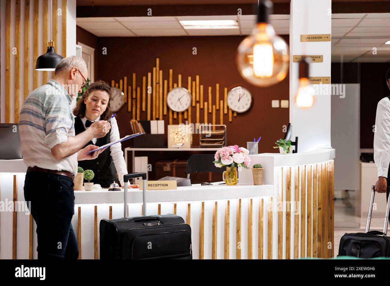 Un touriste masculin senior s'enregistre à la réception de l'hôtel, prêt pour des vacances amusantes. Sympathique concierge femme aide le voyageur homme âgé pendant qu'il remplit les formulaires de réservation. Banque D'Images
