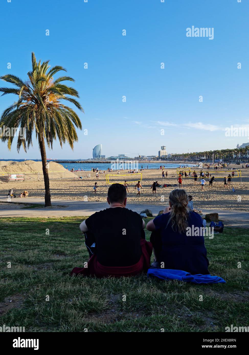 Un couple est assis sur l'herbe près de la plage. En arrière-plan la plage et les gens jouant et se reposant. Barcelone, Catalogne, Espagne. Banque D'Images