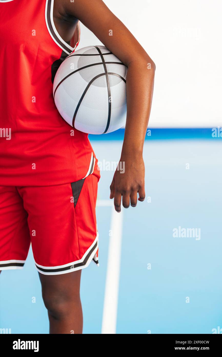 Joueur de basket-ball photo vertical dans un uniforme rouge tenant une balle blanche sur sa hanche, sur un terrain de jeu. Image représentant la passion et le dévouement aux Banque D'Images