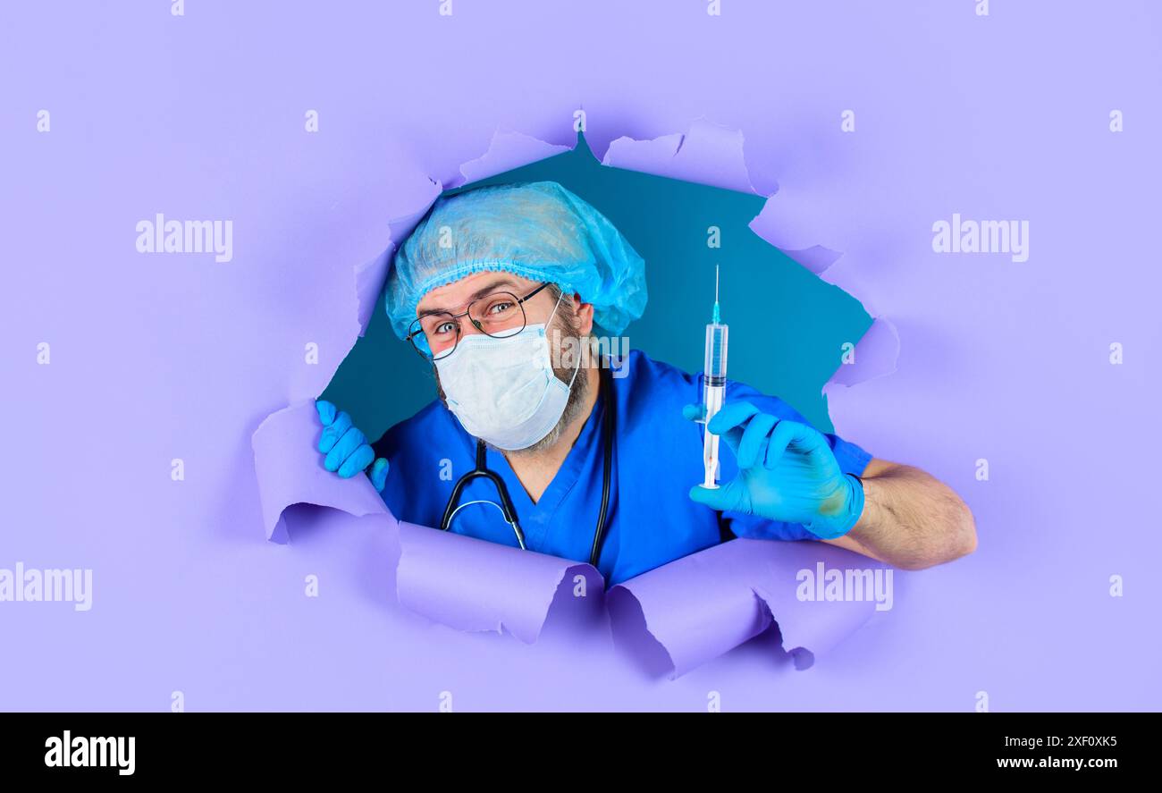 Docteur en manteau médical et gants en caoutchouc avec seringue regardant à travers le trou de papier. Le médecin dans le chapeau chirurgical et le masque médical tiennent la seringue avec le vaccin Banque D'Images