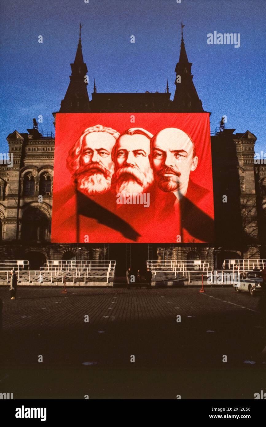 Avril 1988 : une scène de fin de soirée sur la place Rouge à Moscou peu avant les célébrations annuelles du 1er mai. Une femme passe devant une grande bannière rouge, montée sur la façade du grand magasin GUM, représentant les pionniers du communisme russe Karl Marx, Friedrich Engels et Vladimir Lénine. La police reste près des sièges temporaires, leur voiture garée à proximité. Banque D'Images