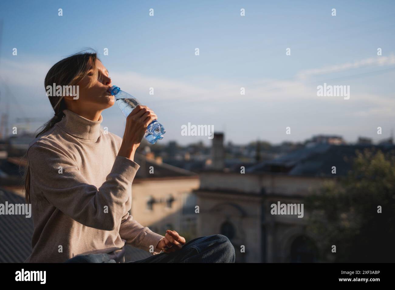 Une femme est assise sur un toit buvant de l'eau. Le ciel est bleu et la femme regarde la ville. Banque D'Images