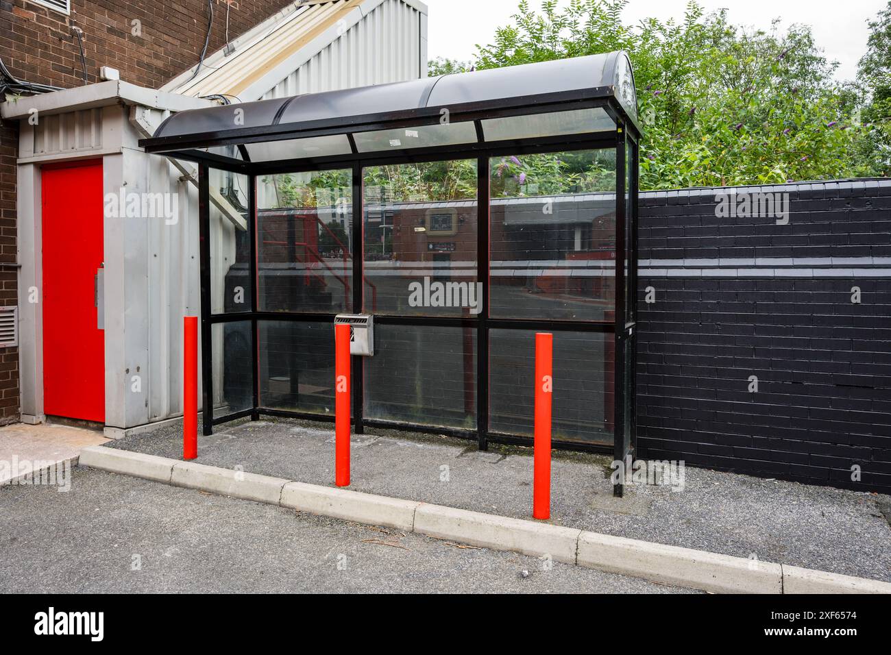 Abri fumeur en verre vide avec des poteaux rouges dans un cadre urbain, entouré de béton et de verdure. Banque D'Images
