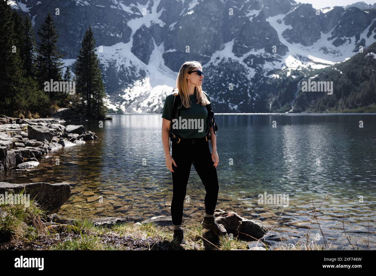 Femme blonde avec sac à dos debout à pleine hauteur par le lac, les montagnes et la neige en toile de fond Banque D'Images