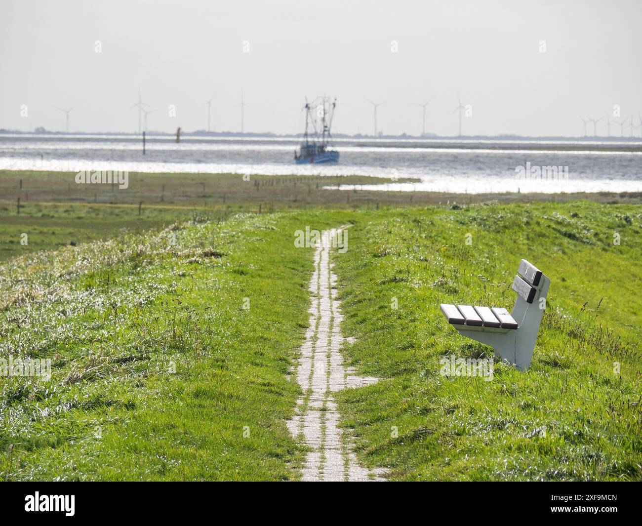 Un chemin étroit avec un banc blanc mène à la mer, où un bateau et des moulins à vent peuvent être vus, Spiekeroog, mer du Nord, allemagne Banque D'Images