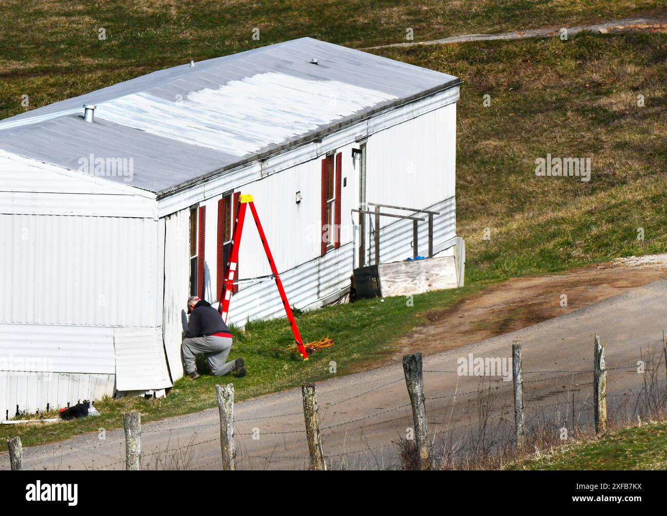 Un homme âgé répare la jupe autour de son mobil-home. La maison est blanche avec un toit argenté. Il porte une casquette et un échauffement, un pantalon gris. Banque D'Images