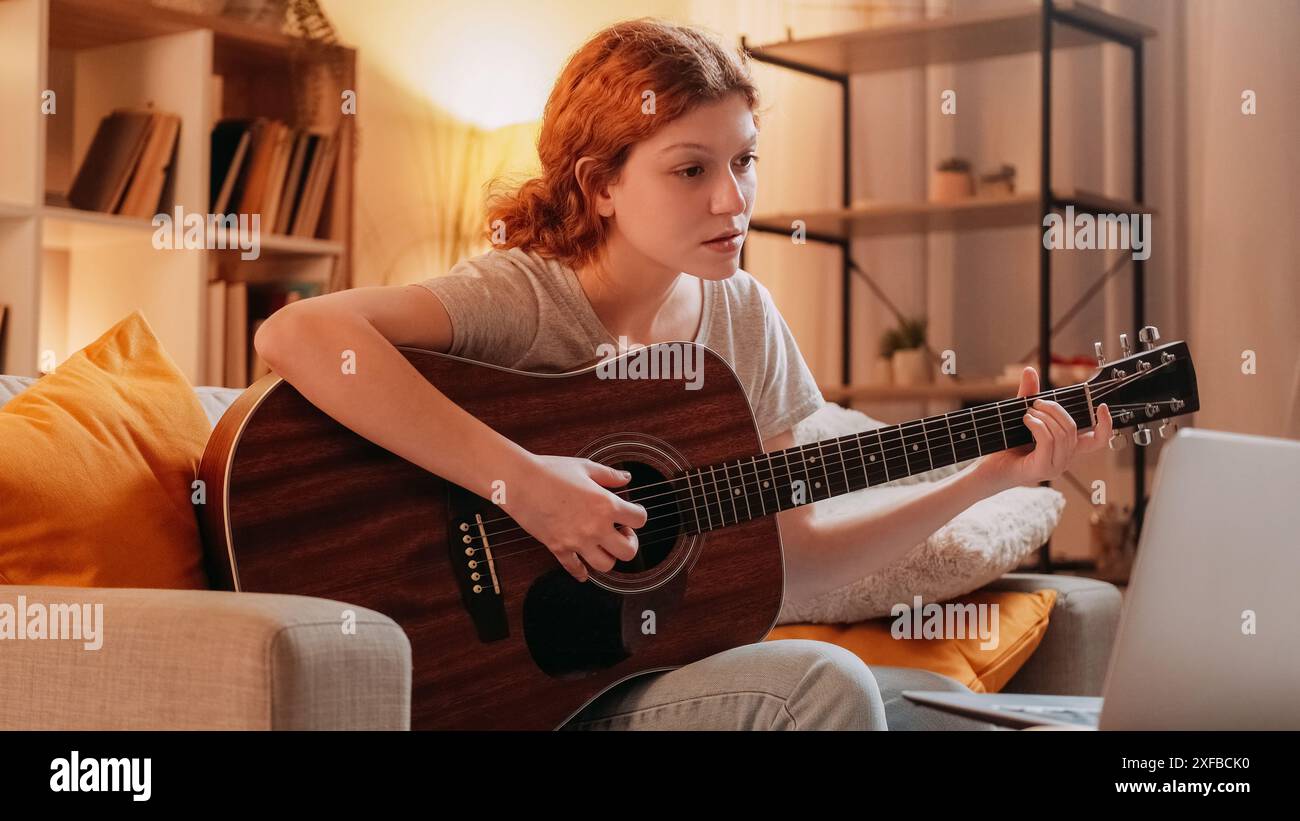 Apprentissage de la guitare. Cours de musique sur Internet. Femme curieuse inspirée jouant un instrument acoustique regardant une leçon vidéo sur ordinateur portable à la maison. Banque D'Images