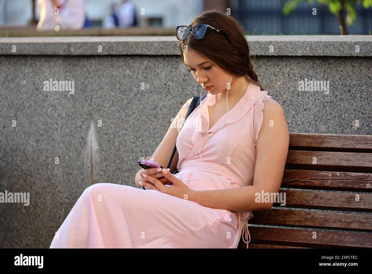 Fille attrayante assise sur le banc de rue avec smartphone dans les mains, en utilisant le téléphone portable dans la ville d'été Banque D'Images