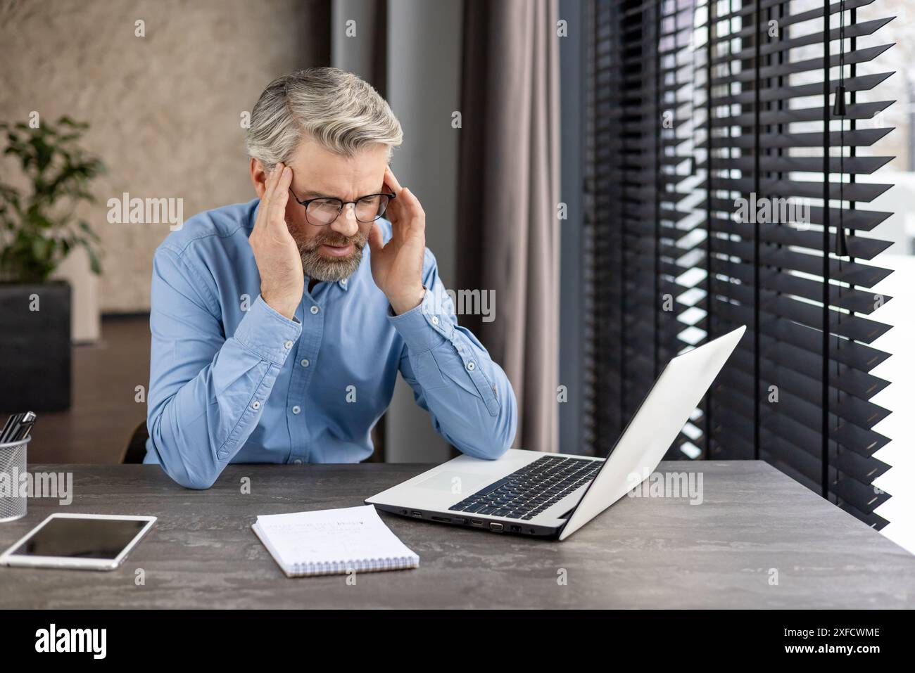 Un employé de bureau vêtu d'une chemise bleue et de lunettes est assis à un bureau et semble frustré. L'homme a les mains sur ses tempes, ce qui indique un stress ou des maux de tête. Bloc-notes, tablette et ordinateur portable sont sur le bureau. Banque D'Images