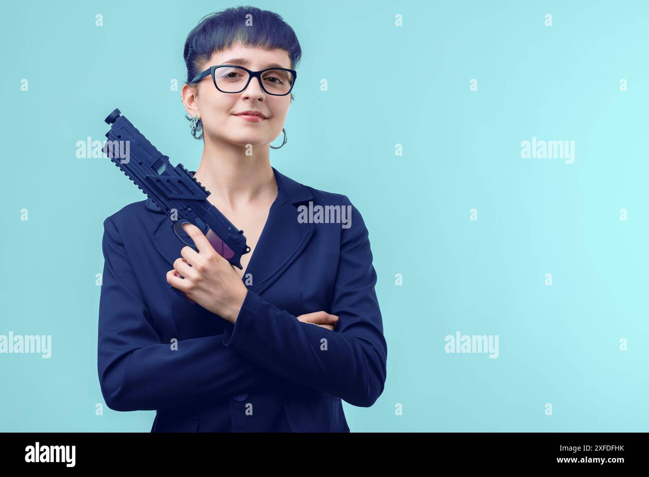 Femme d'affaires tient un pistolet tout en se tenant debout avec ses bras croisés sur un fond bleu. L'image évoque les concepts de pouvoir, de sécurité et de défense Banque D'Images