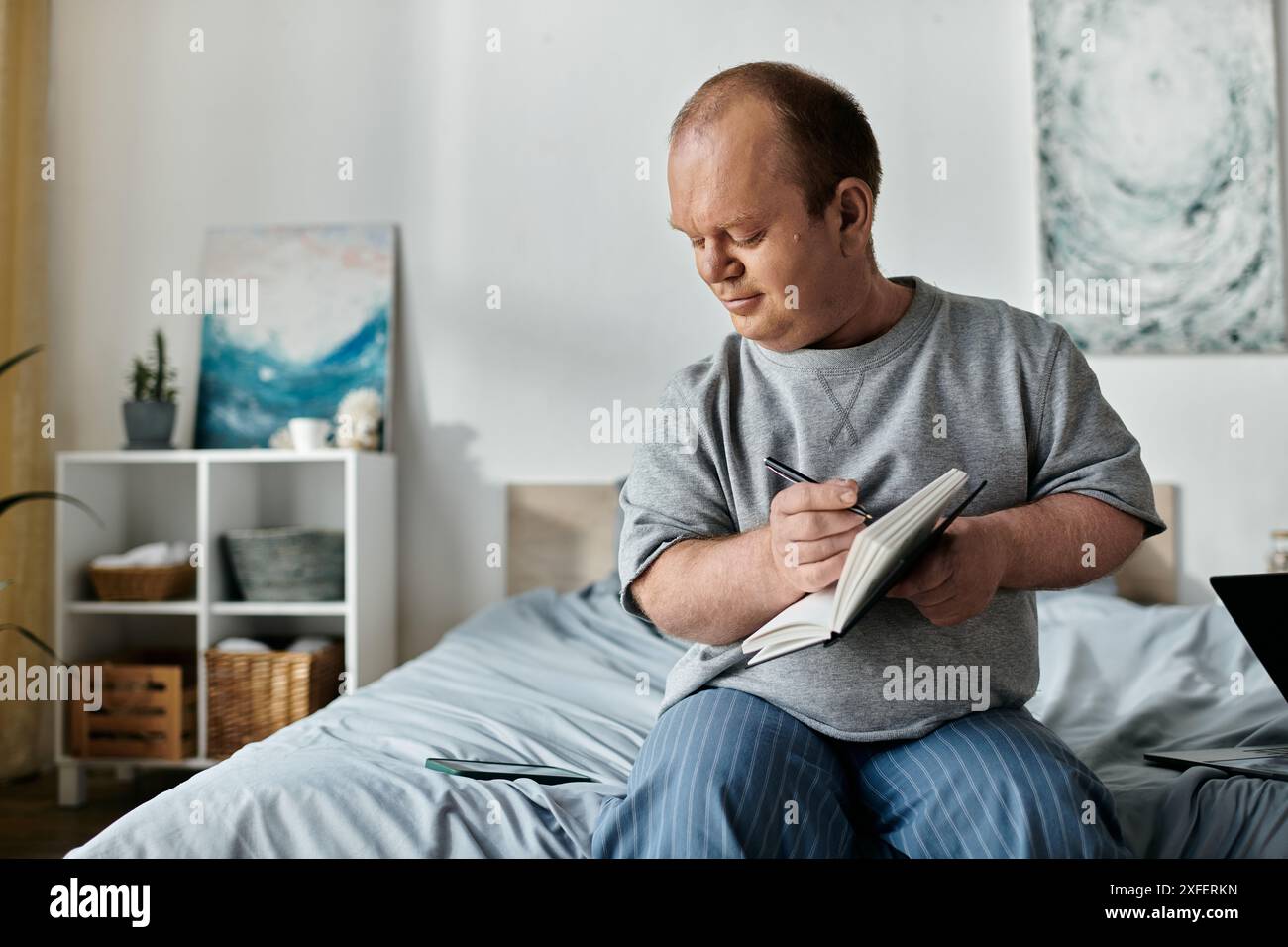 Un homme avec inclusivité est assis sur un lit, écrivant soigneusement dans un cahier. Banque D'Images