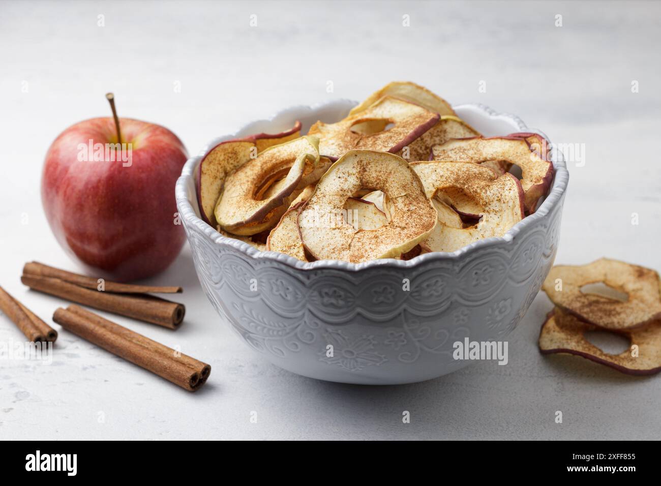 Pommes séchées maison à la cannelle, chips de pomme dans un bol avec des bâtons de cannelle sur un fond gris clair. Délicieux en-cas sain Banque D'Images