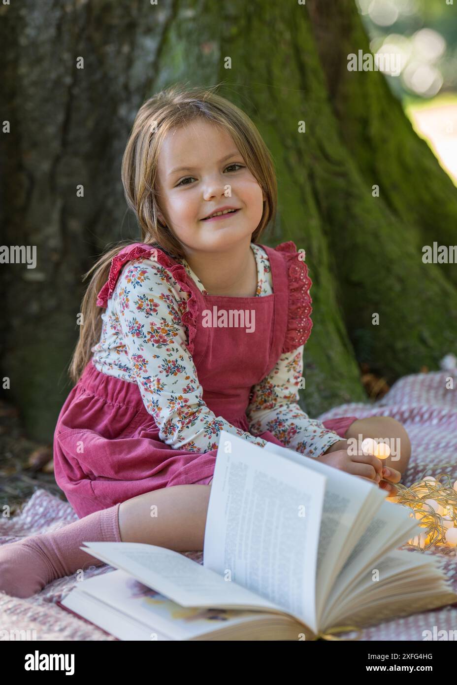 Une petite fille de 5 ans est assise près d'un grand arbre dans le parc. Sourit et regarde la caméra. A proximité se trouve un livre ouvert et des lanternes. Banque D'Images