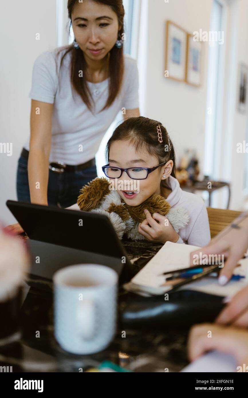 Femme à côté de la fille étudiant à travers la tablette numérique à la maison Banque D'Images