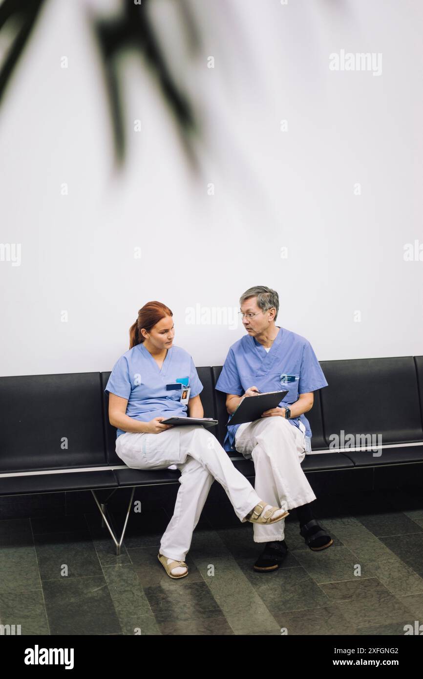 Collègues médicaux masculins et féminins discutant au-dessus de documents tout en étant assis sur une chaise près du mur à l'hôpital Banque D'Images