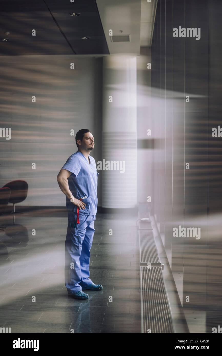 Pleine longueur du médecin masculin debout avec les bras akimbo près de la fenêtre à l'hôpital Banque D'Images