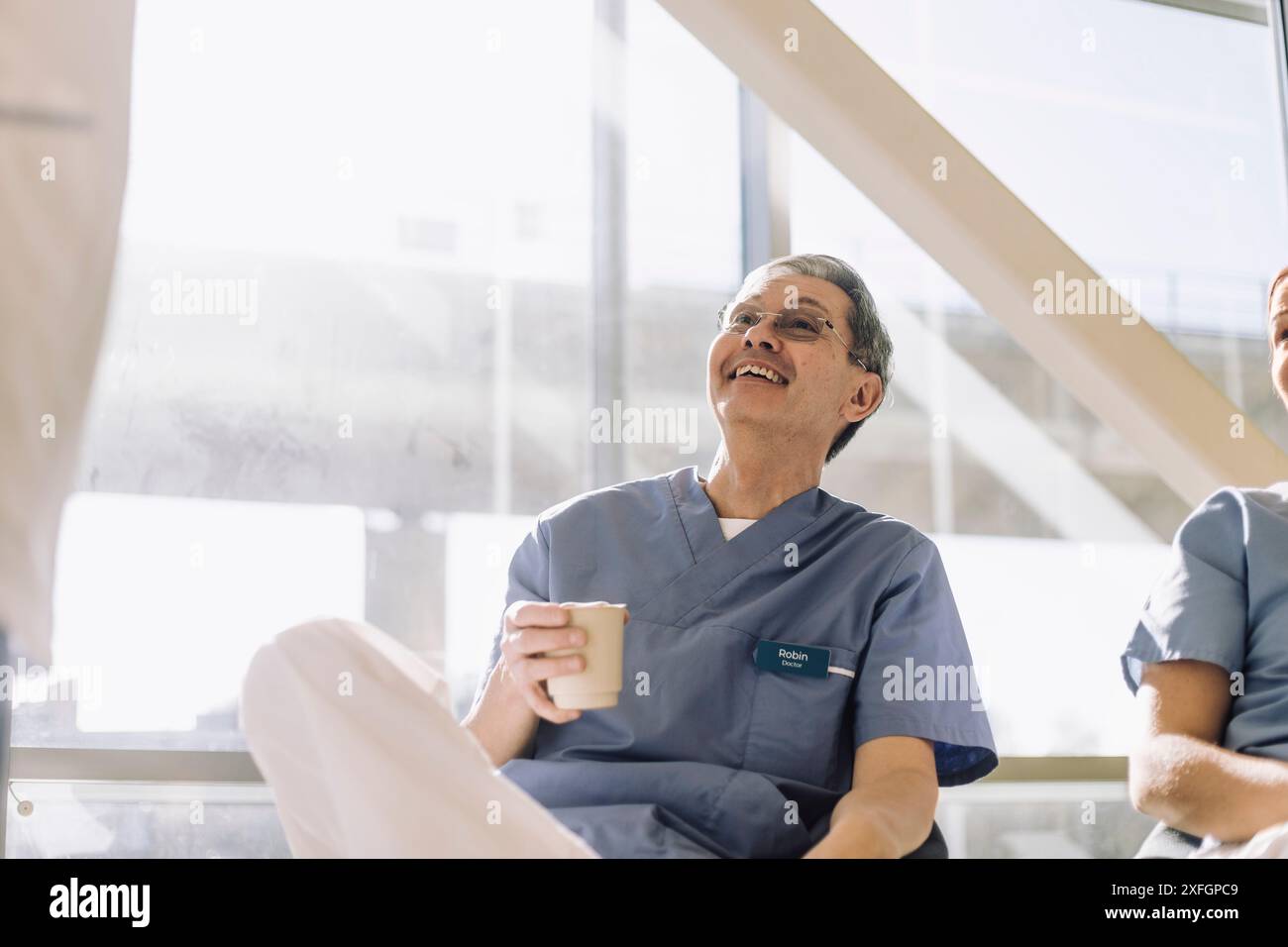 Souriant expert médical masculin senior tenant un verre à café tout en étant assis à la cafétéria avec des collègues Banque D'Images