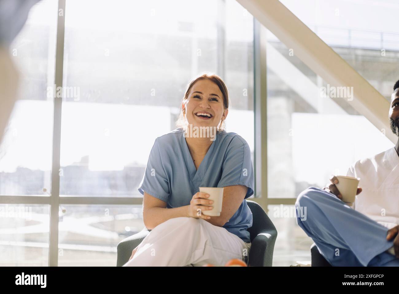 Expert médical féminin souriant tenant une tasse jetable tout en étant assis avec un collègue à la cafétéria Banque D'Images