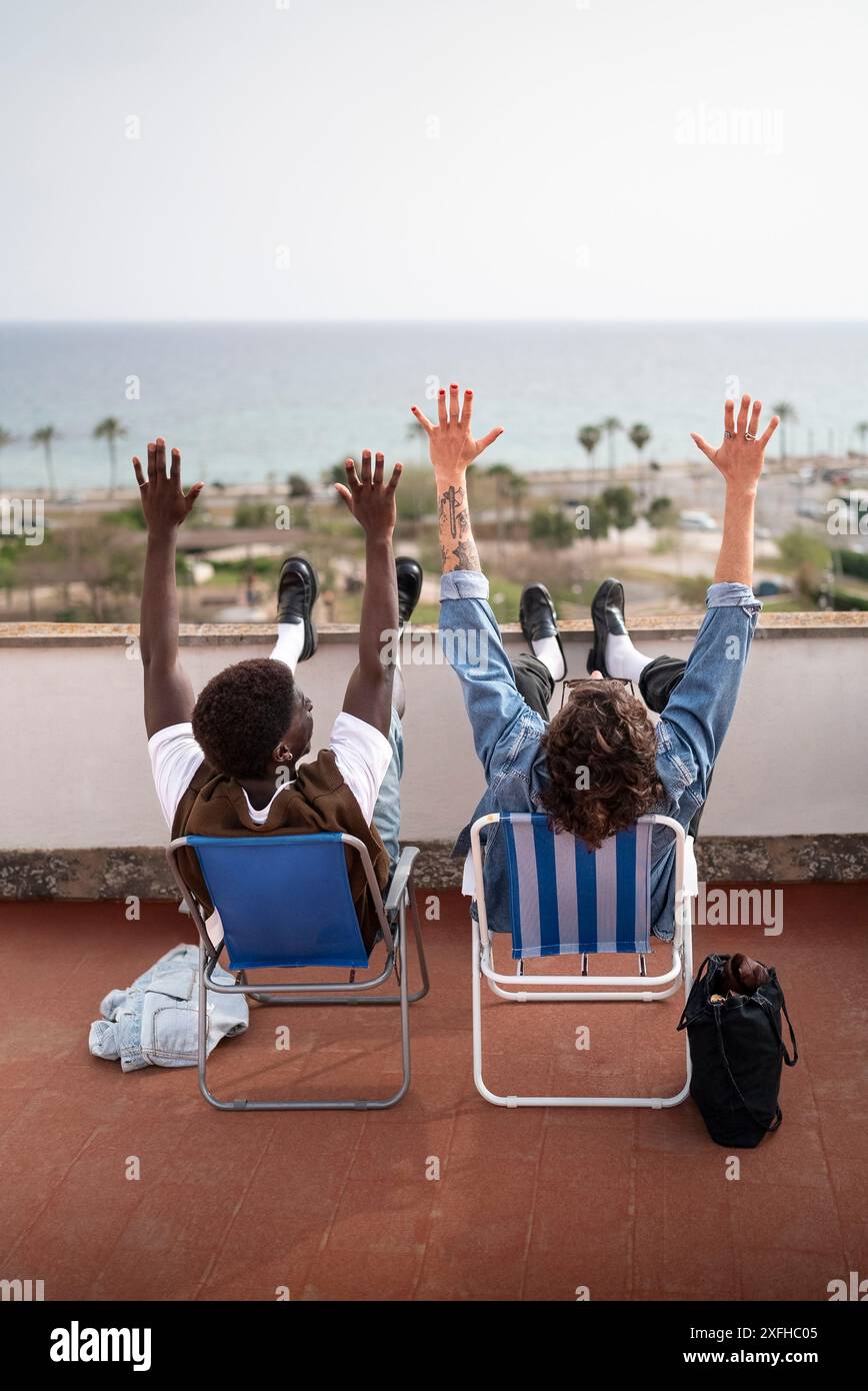 Vue arrière d'amis masculins insouciants assis avec les bras levés sur des chaises à la terrasse du bâtiment Banque D'Images