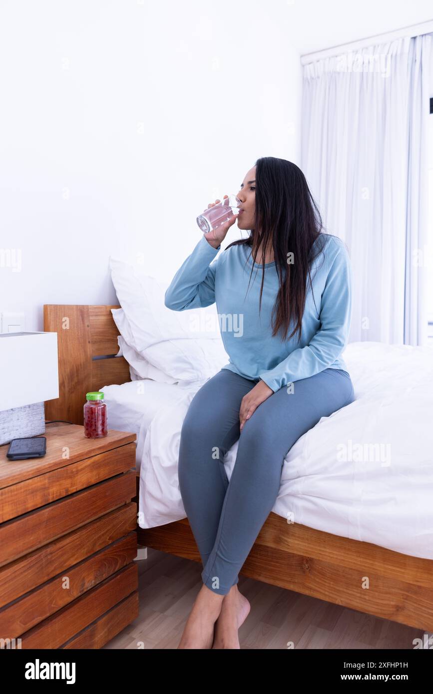 Eau potable, femme assise sur le lit dans une chambre confortable, routine matinale relaxante Banque D'Images