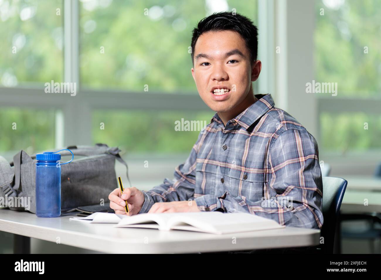 Un jeune adolescent est assis à une table de classe avec un livre, des notes et une bouteille d'eau pour étudier et apprendre, souriant à la caméra Banque D'Images