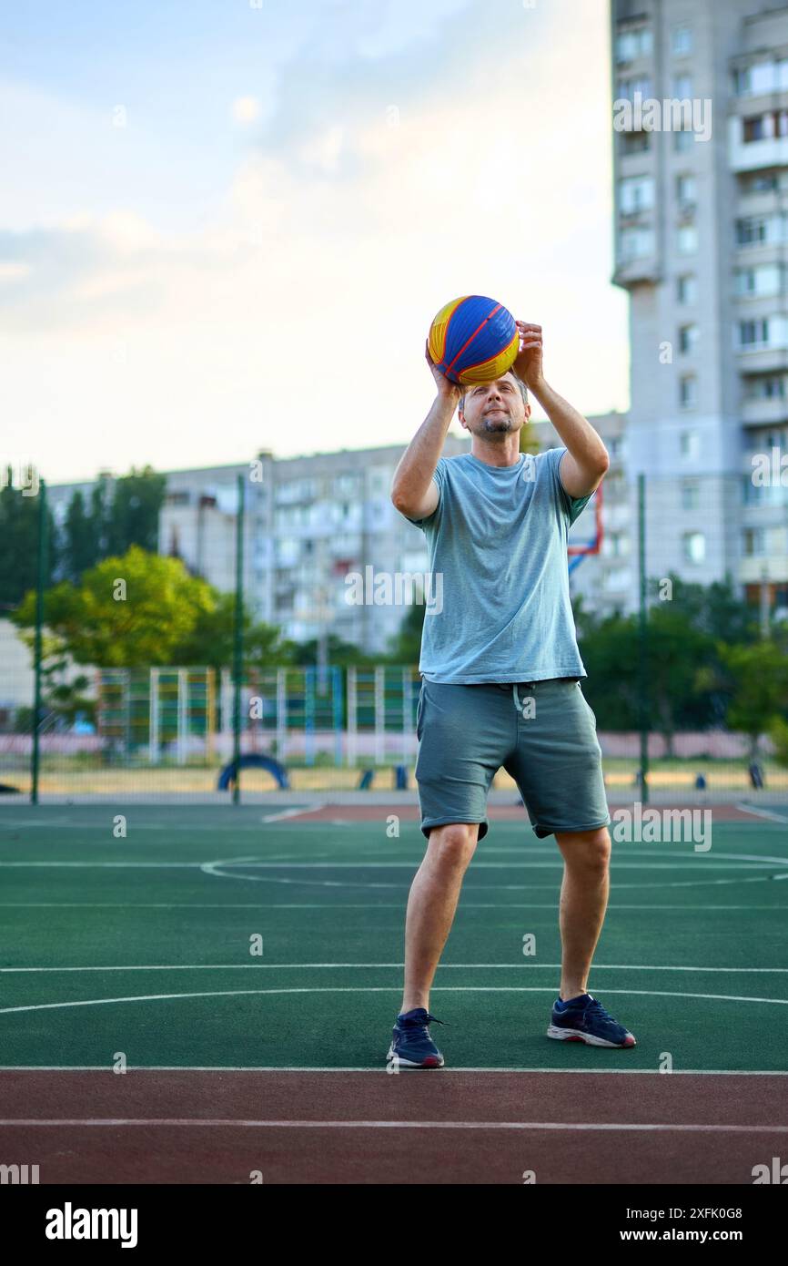 Homme d'âge moyen jouant au basket-ball sur le court de la ville le jour ensoleillé. Homme fort faisant du sport dehors. Bâtiments résidentiels et arbres en arrière-plan. SpO Banque D'Images