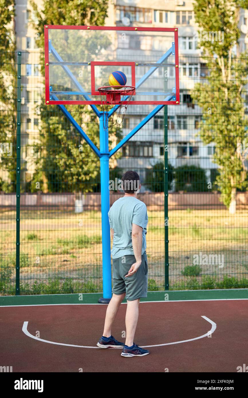 Homme d'âge moyen jouant au basket-ball sur le court de la ville le jour ensoleillé. Homme fort faisant du sport dehors. Bâtiments résidentiels et arbres en arrière-plan. SpO Banque D'Images