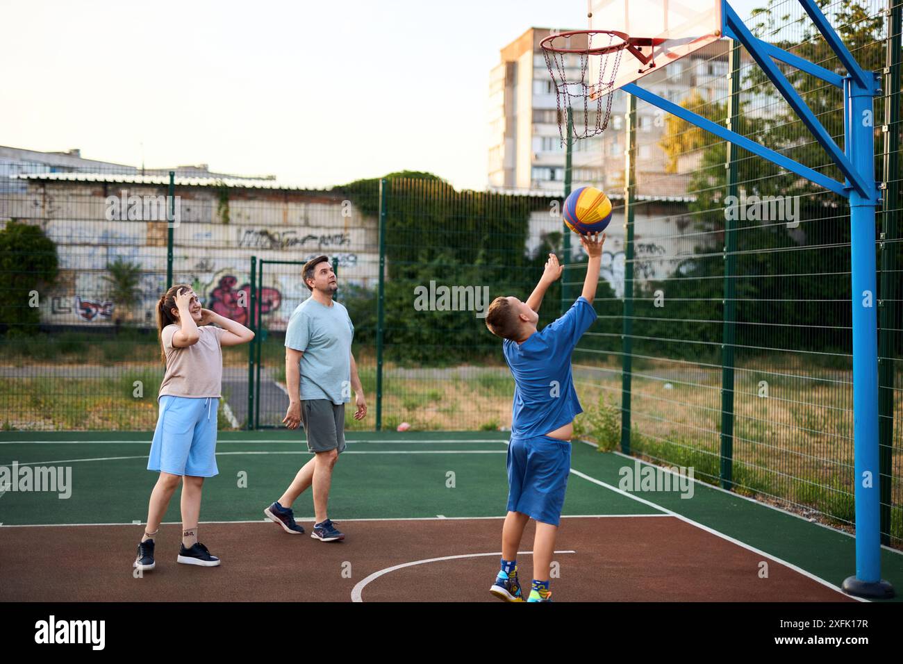 Motion shot d'un père heureux et d'enfants jouant au basket-ball ensemble à l'extérieur sur un terrain de basket-ball moderne parmi des maisons ordinaires, dans une zone urbaine Banque D'Images