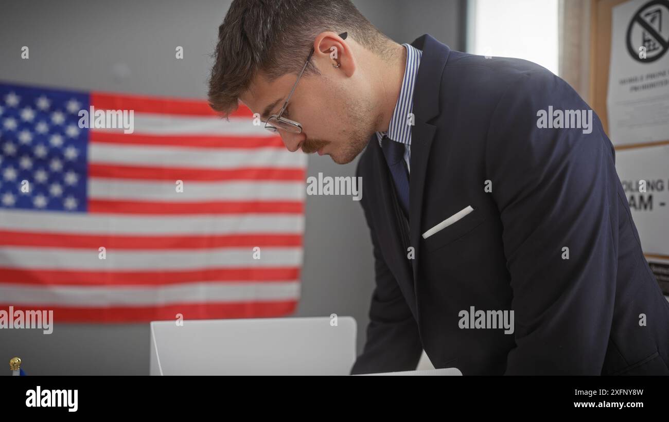 Un jeune homme d'affaires hispanique avec des lunettes et une moustache travaille sur un ordinateur portable dans un bureau à thème patriotique avec un drapeau américain. Banque D'Images