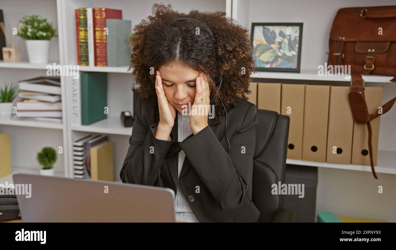 Une jeune femme hispanique aux cheveux bouclés éprouvant du stress sur son lieu de travail, portant des écouteurs et un costume. Banque D'Images
