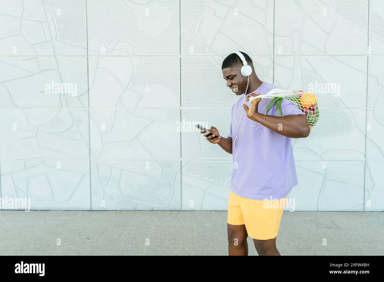 Mode de vie écologique (zéro déchet) et concept de régime alimentaire sain. Homme afro-américain porter des écouteurs en vêtements de sport à l'aide d'un smartphone tenant maille réutilisable Banque D'Images